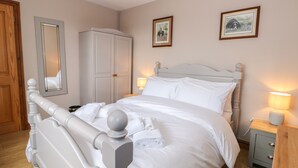 A neatly made bed with white linens, a wooden headboard, and a bedside table with a lamp. There is a wardrobe with a mirror above it and two framed pictures on the wall.