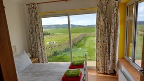 A bedroom with a large bed, a view of a green field through a window, and a wooden floor. - Orchard Chalet (Liskeard)