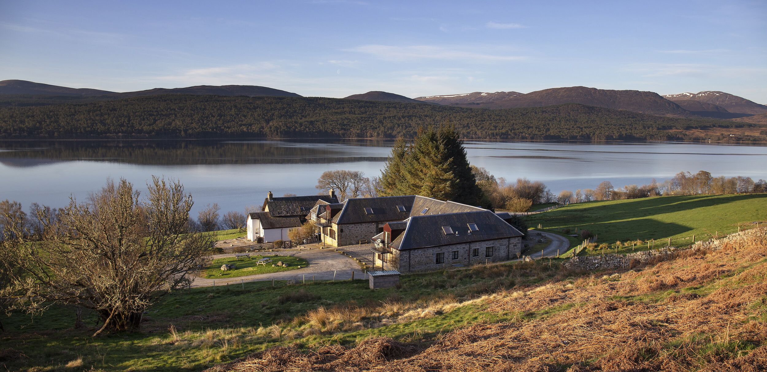 Une propriété au bord du lac, avec des bâtiments en pierre, une pelouse verdoyante et une vue sur le lac et les montagnes.