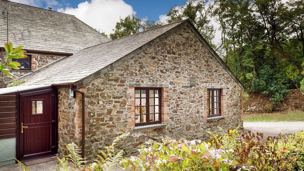 Une maison en pierre avec une porte et des fenêtres de couleur brun foncé, entourée d’une végétation luxuriante.