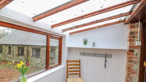 Une pièce comprenant une chaise en bois, une étagère avec des crochets, une fenêtre donnant sur un bâtiment en pierre et un vase rempli de fleurs.