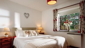 Une chambre à coucher bien rangée, avec un lit, deux tables de chevet dotées de lampes, un cœur décoratif accroché au mur, et une fenêtre avec des rideaux et un vase de fleurs.