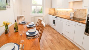 A modern kitchen with white cabinets, a wooden dining table set for four, and a built-in washing machine. - Daisy Cottage (Leyburn)