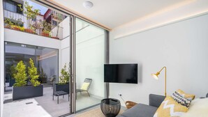A modern living room with a large sliding glass door leading to an outdoor balcony, a flat-screen TV mounted on the wall, a grey sofa with patterned cushions, and a floor lamp. - WOW! Urban Retreat - Heated Pool, Daily Breakfast & Housekeeping    (Porto)