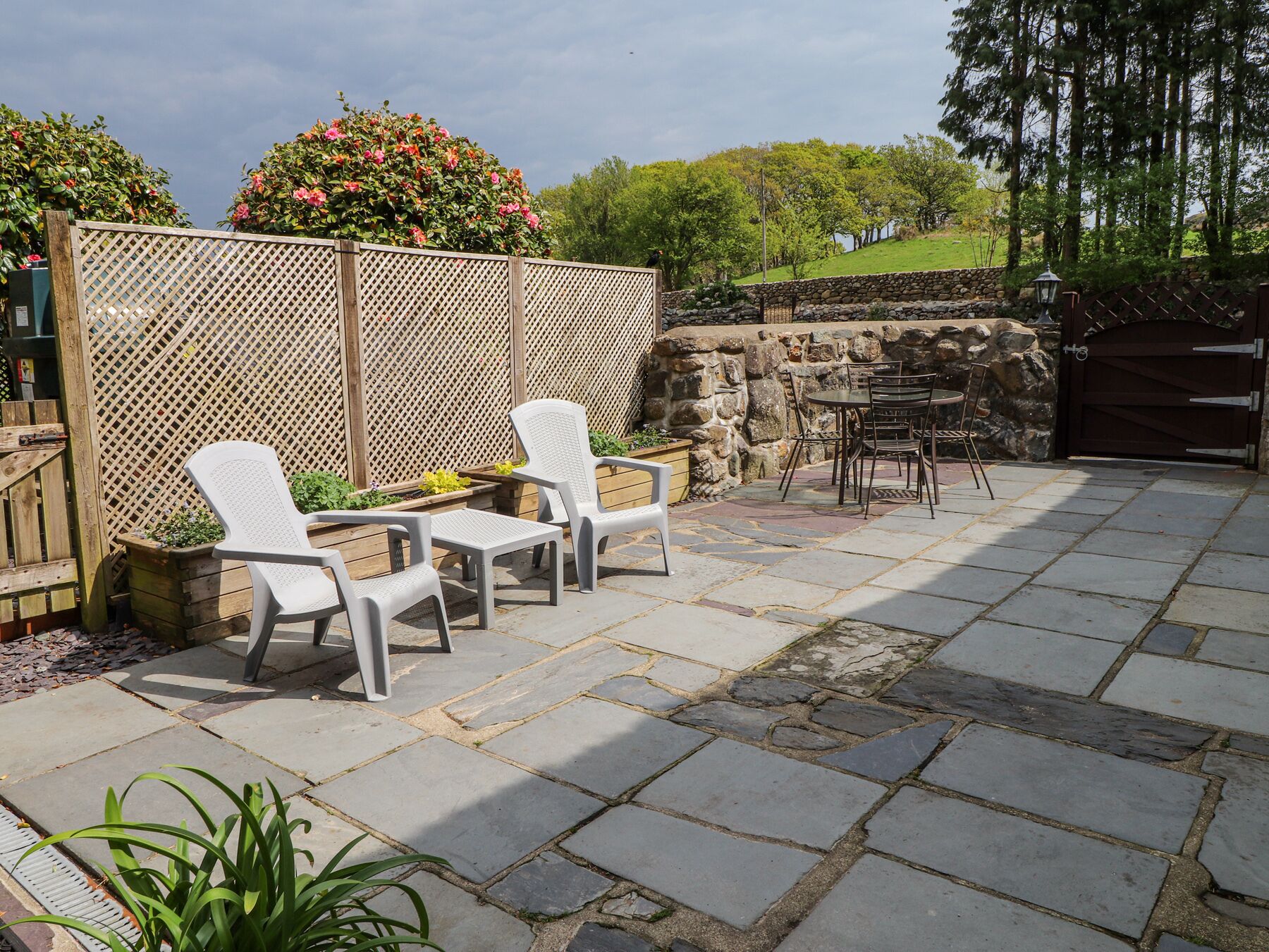 Un patio exterior con suelo de piedra, sillas de plástico blanco, una mesa y una valla de madera.