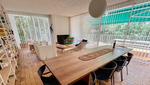 Ein geräumiger Essbereich mit einem langen Holztisch, schwarzen Stühlen und einer dekorativen Tischläufer. Links befindet sich ein Bücherregal und im Hintergrund ein Sitzbereich mit Fernseher und bunten Stühlen.