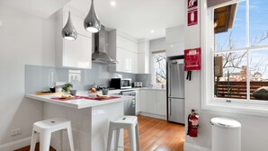 Une cuisine moderne avec des meubles blancs, des appareils en acier inoxydable et un îlot central doté de tabourets. Des luminaires suspendus sont placés au-dessus de l’îlot, un extincteur est fixé au mur, et une fenêtre offre une vue sur l’extérieur.