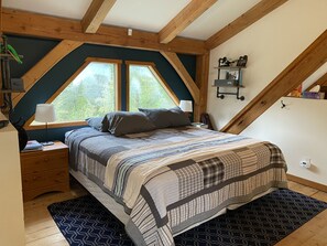 A bedroom with a sloped ceiling, a large bed with a patterned bedspread, two windows, a nightstand, a lamp, and a small shelf with items. - Moyie Lake Cabin (Moyie)