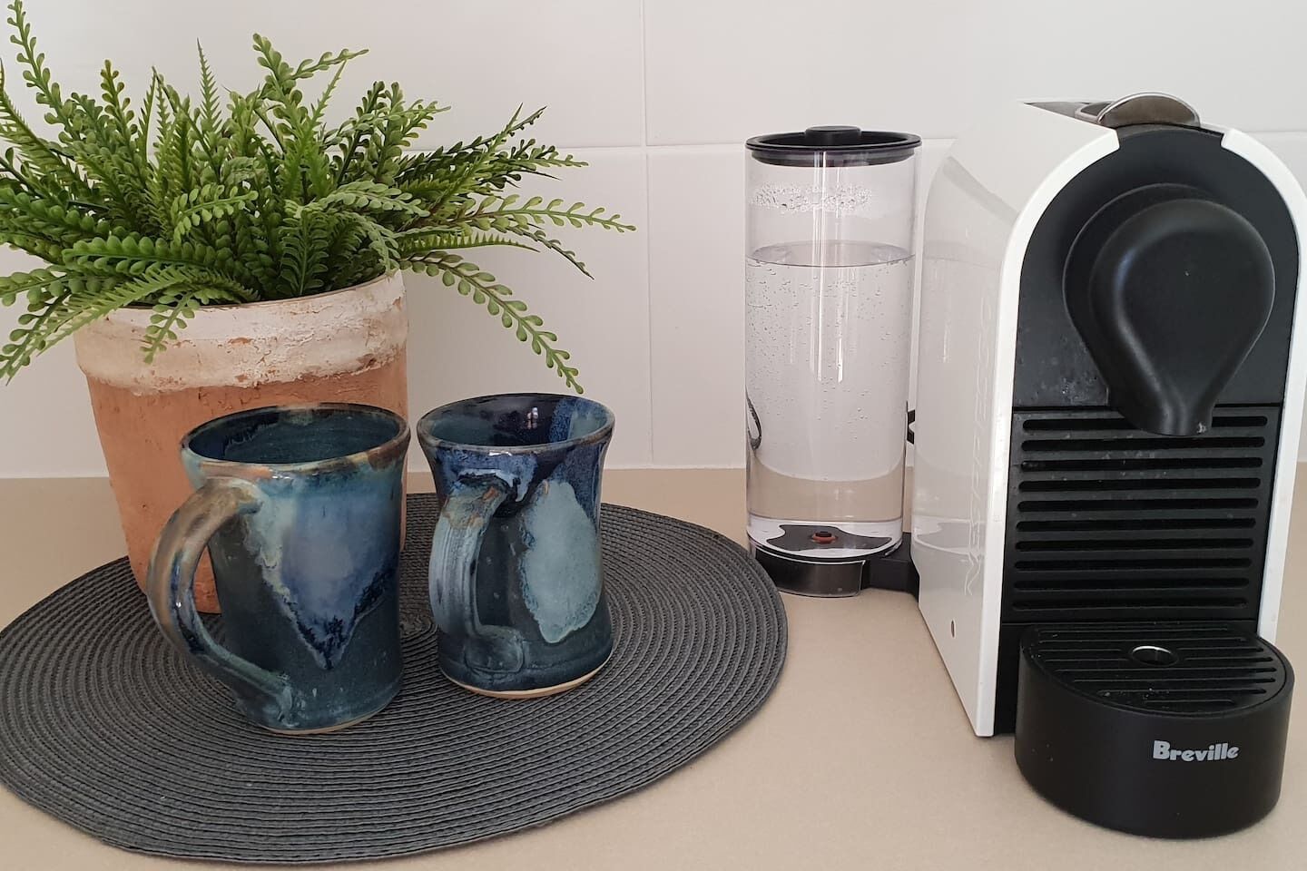 Une machine à café de la marque Breville, deux tasses en céramique et une fougère en pot, le tout posé sur un tapis tissé.