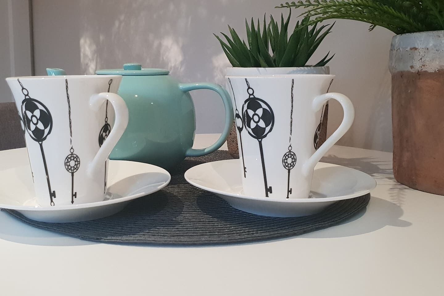 Two white cups with black key designs on a blue mat, a teal teapot, and a green plant in a pot.
