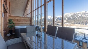 Eine Holzblockhütte mit einem Glastisch und Stühlen, einem Whirlpool und Blick auf schneebedeckte Berge.