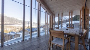 Eine Holzterrasse mit Tisch und Stühlen, große Fenster mit Blick auf schneebedeckte Gebäude und Berge.