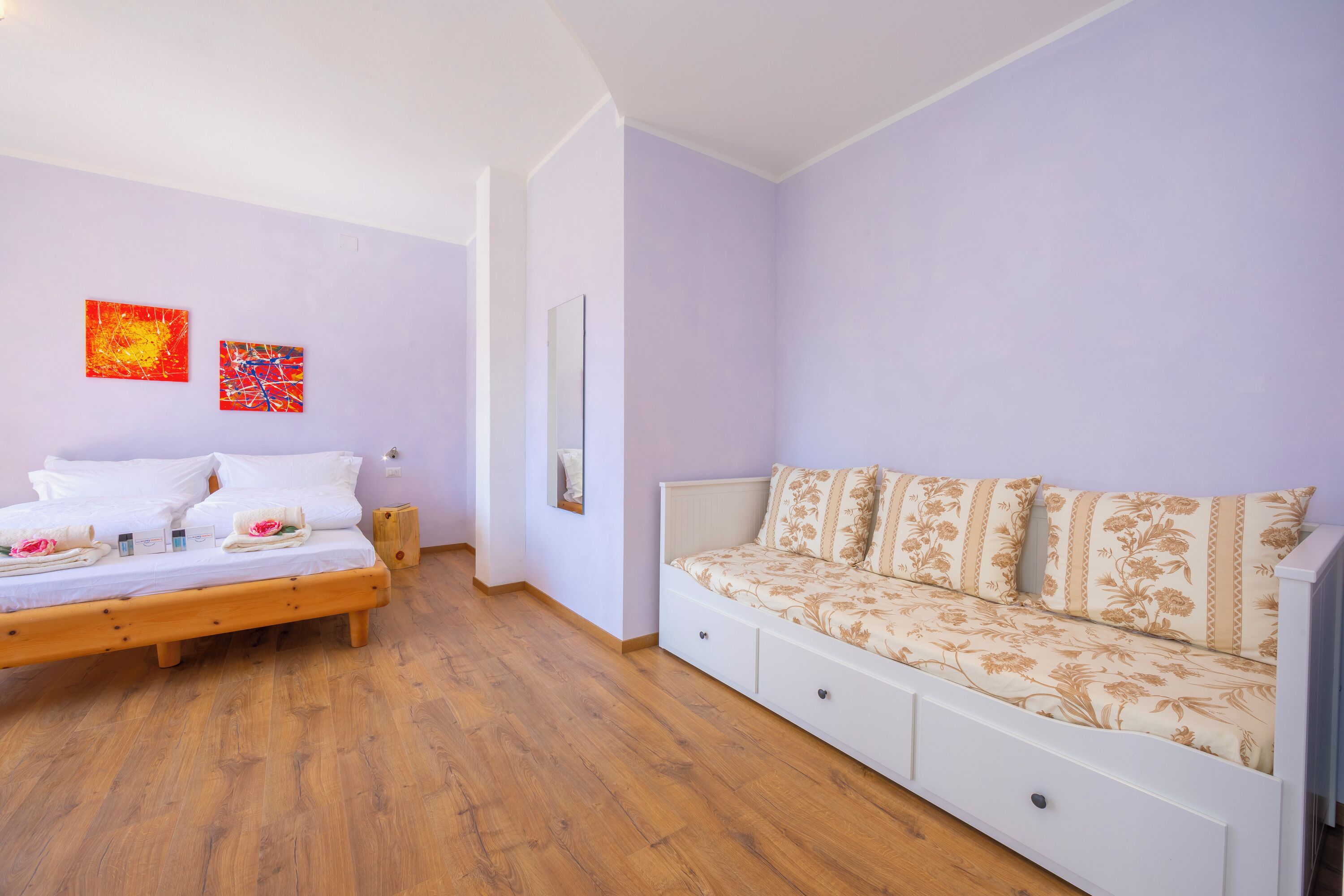 A bedroom with a bed, a sofa, wooden flooring, and two wall art pieces.