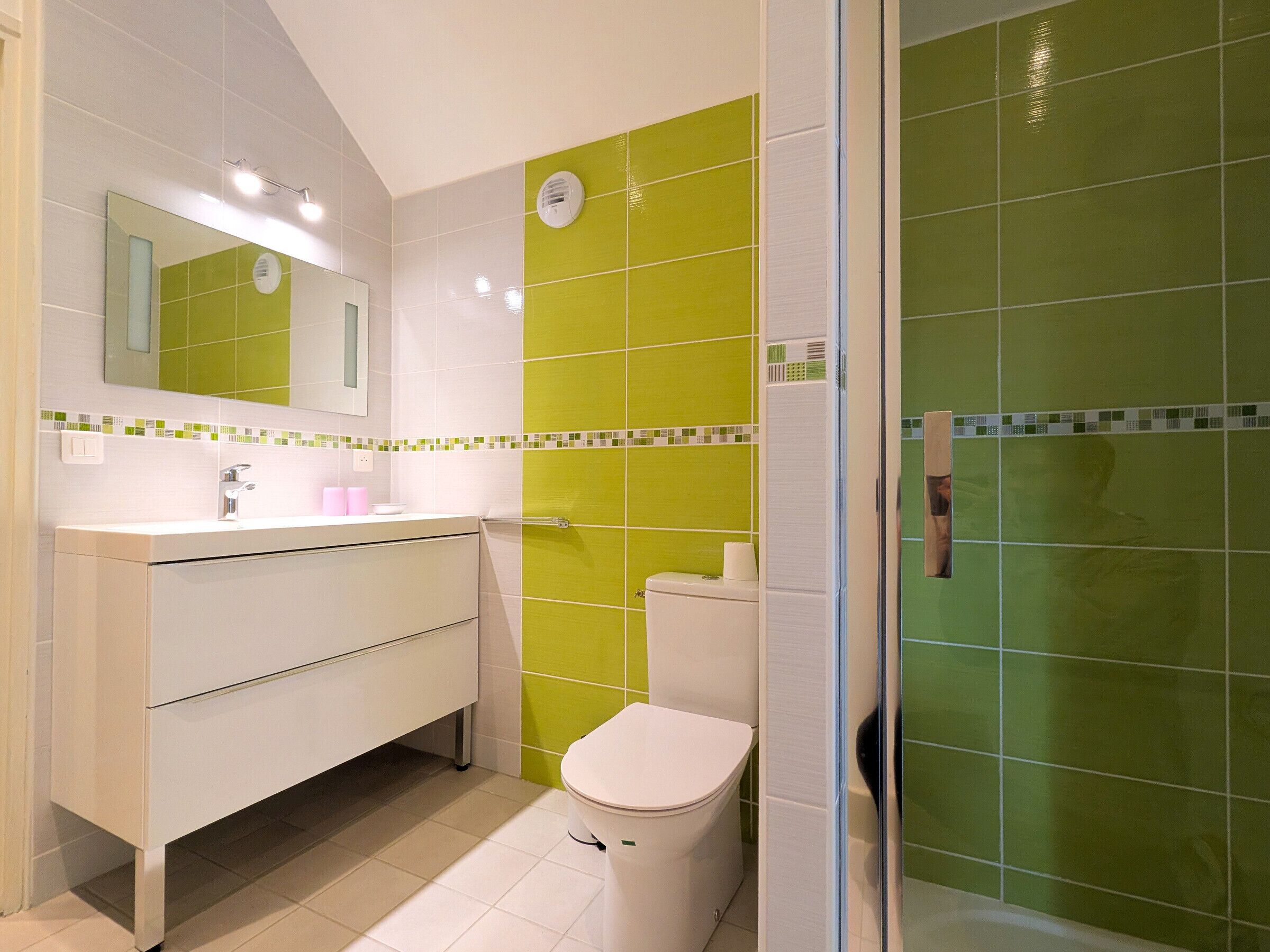A bathroom with a white sink, mirror, and toilet, and a shower area with green tiles.