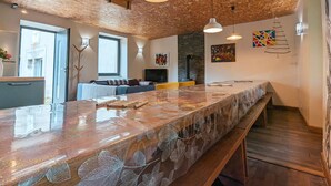 A modern kitchen with a wooden ceiling, a long island with a glass top, and a dining area with a sofa and yellow chairs. - Charming Village House with Shared Heated Pool, Near Attractions, and Private Garden (Le Menoux)