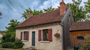 A rustic two-story house with a red-tiled roof, a wooden side extension, and a small front porch. - Charming Village House with Shared Heated Pool, Near Attractions, and Private Garden (Le Menoux)