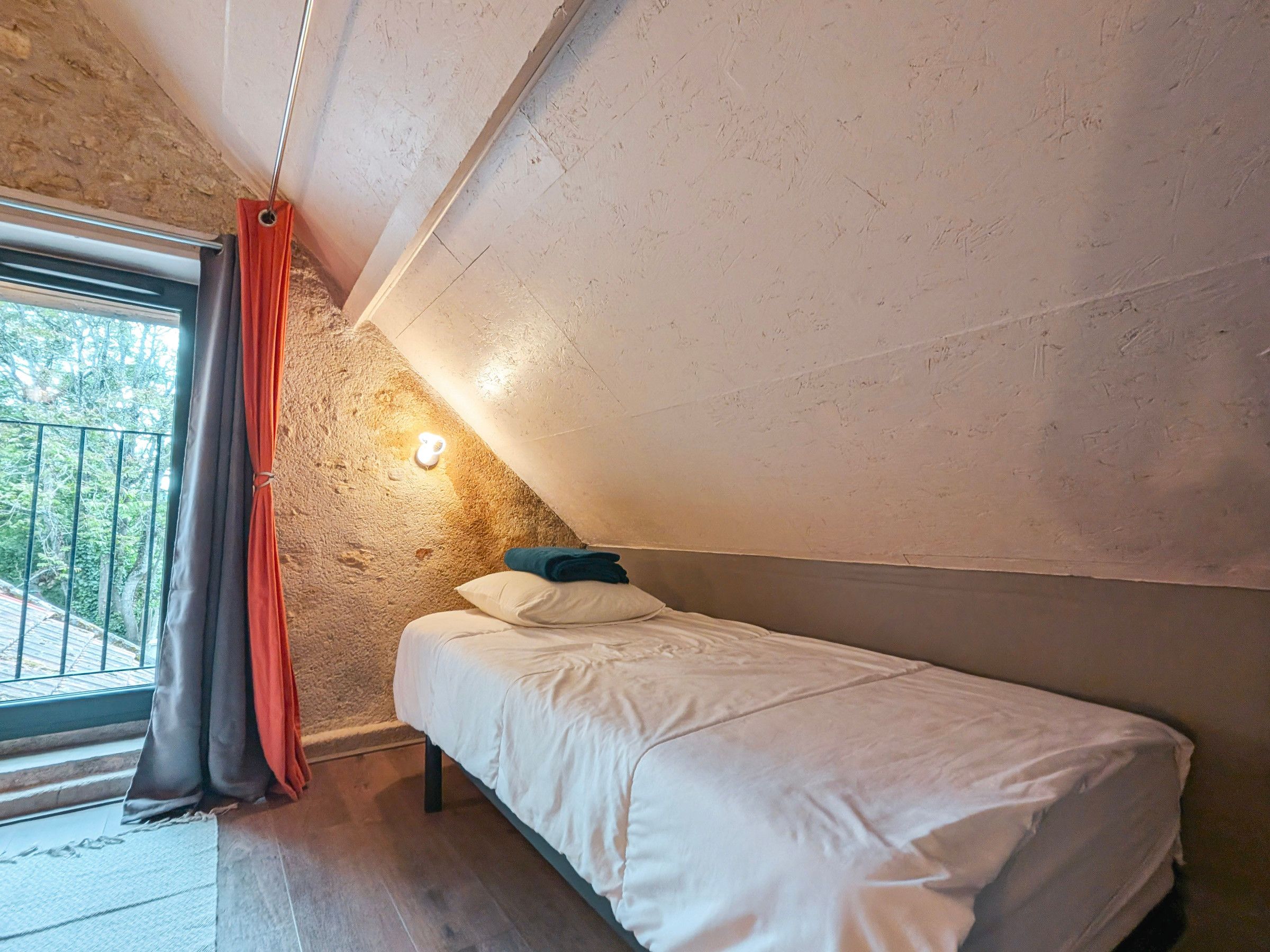 A small, single-bed room with a sloped ceiling, a window with curtains, and a wall-mounted light.