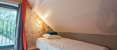 A small, single-bed room with a sloped ceiling, a window with curtains, and a wall-mounted light.