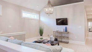 A modern living room with a sofa set, a dining table, a chandelier, a TV mounted on the wall, and a small plant on a side table.