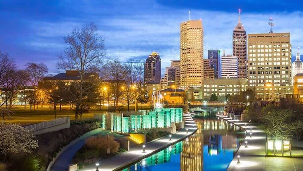 A cityscape with a prominent building in the background, a well-lit pathway leading to a body of water, and a park area with trees and a fountain.