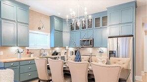 A modern kitchen with teal cabinetry, a chandelier, and a dining area with beige upholstered chairs.