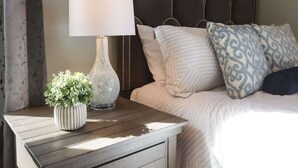 Een net opgemaakt bed met gekleurde kussens, een bedlampje en een kleine plant in een potje op een houten nachtkastje.