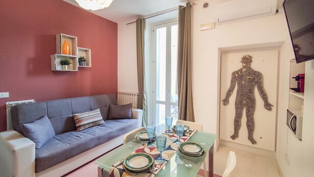 A living room with a sofa, a dining table set with plates and glasses, a wall-mounted shelf with decorative items, and a large wall art piece. - Casa Chiara in centro con corte privata (San Benedetto del Tronto)