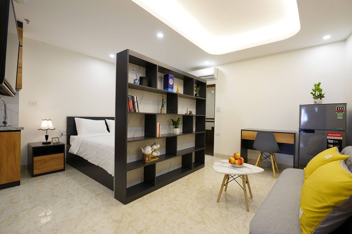 A modern hotel room with a bed, a bookshelf, a small table with fruit, a sofa with yellow cushions, and a refrigerator.