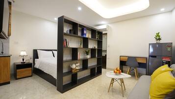 A modern hotel room with a bed, a bookshelf, a small table with fruit, a sofa with yellow cushions, and a refrigerator.