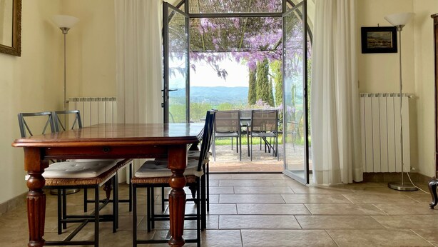 Un coin repas avec une table et des chaises en bois, une vue sur une terrasse extérieure accessible par un passage en arc, où se trouvent une table et des chaises, et un radiateur fixé au mur.