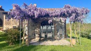 Une pergola en pierre ornée de fleurs violettes, avec des sièges d’extérieur, et une vue sur un bâtiment en arrière-plan.