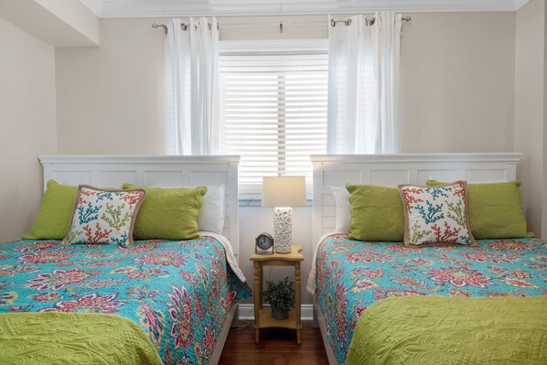 Un dormitorio con dos camas, cada una con ropa de cama verde y almohadones decorativos, una mesita de noche con lámpara y una ventana con cortinas blancas.