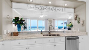 A kitchen with white cabinets, a stainless steel sink, and a dishwasher. There is a large window with a view of the ocean.