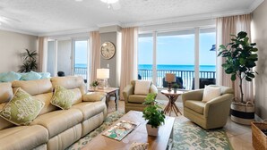 A spacious living room with a beige sofa, two armchairs, a coffee table, and a view of the ocean through large windows.