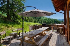An outdoor wooden deck with picnic tables and benches, a barbecue grill, and a large umbrella providing shade.