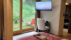 A bedroom with a large window, a bed with a red and white patterned bedspread, a white chair, a desk with a computer, and a television mounted on the wall.