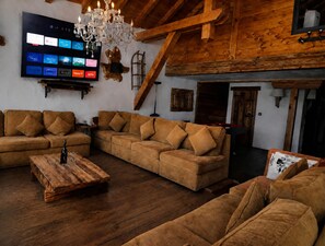 A cozy living room with wooden furniture, a flat-screen TV mounted on the wall, and a chandelier.