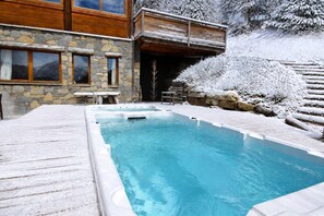 A swimming pool with a wooden deck, stone walls, and a snow-covered landscape.