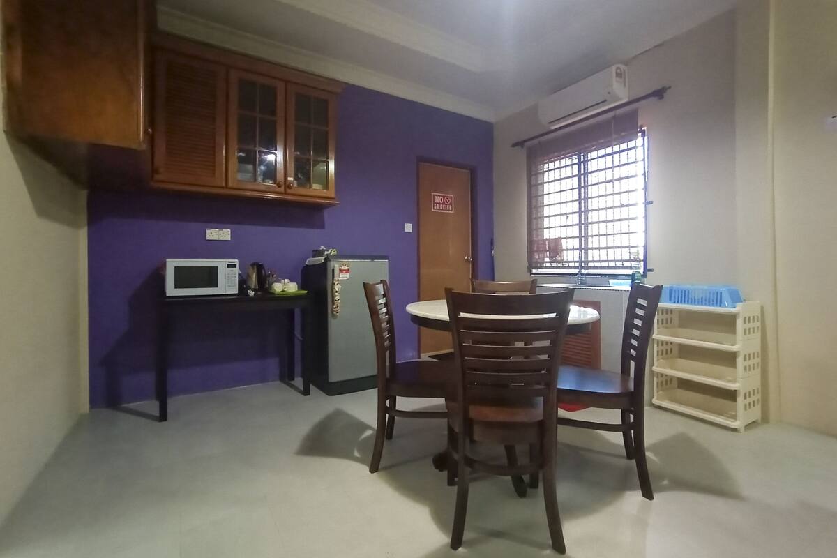 Een kleine keuken met een magnetron, koelkast en houten keukenkastjes. Een eettafel met stoelen en een raam met roedeversiering.