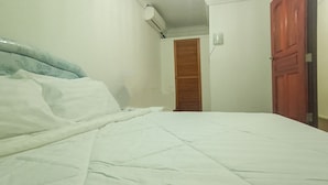 Una cama con cubrecama blanco, un armario de madera y una puerta cerrada en una habitación con paredes blancas.