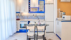 A compact kitchen with white and blue cabinets, a built-in oven, a refrigerator, and a small dining area with two chairs. - Seafront Paradise Home (Skala Fourkas, Halkidiki)