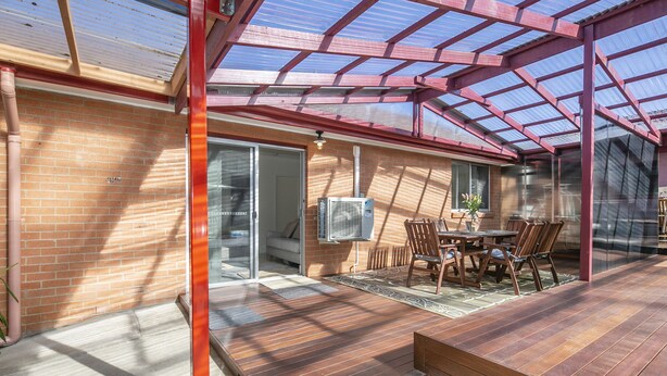 A covered patio with a wooden deck, a glass door leading to a room, and a table with chairs.