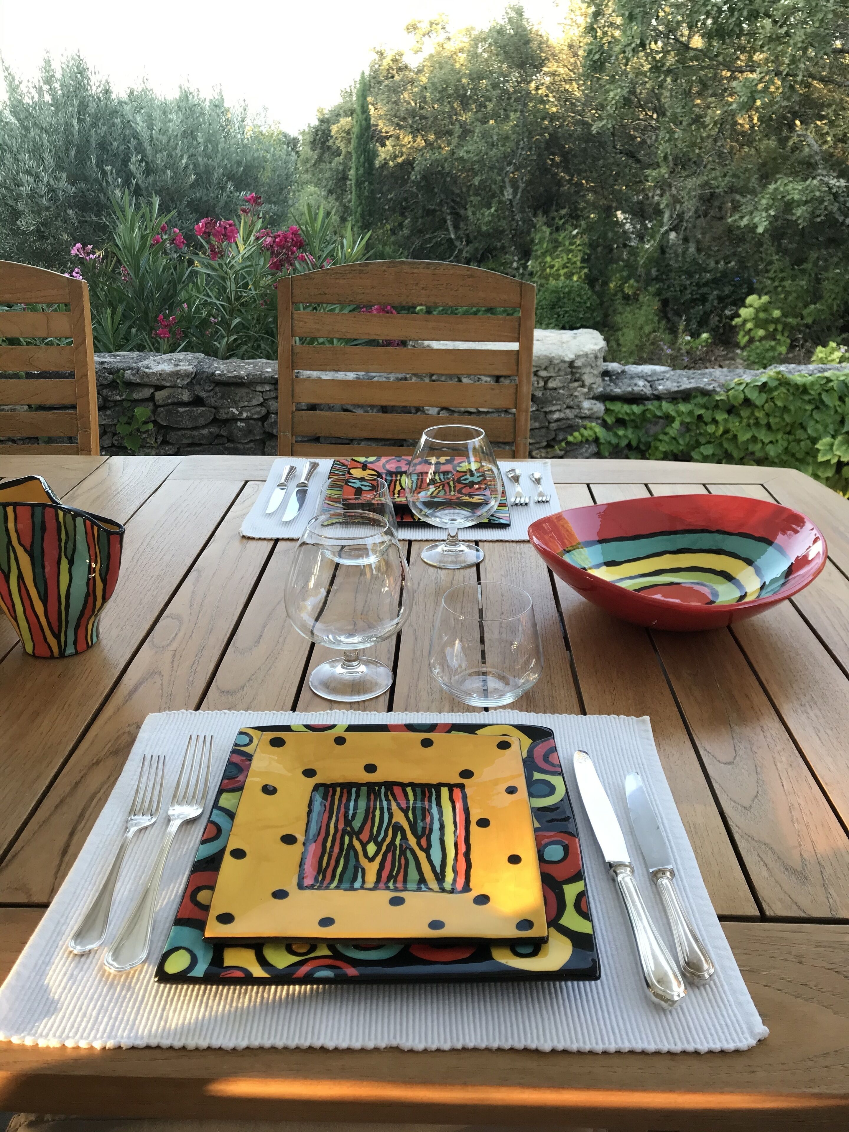 A wooden table set with colorful tableware and cutlery, surrounded by stone walls and greenery.