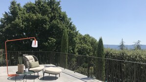 En balkon med to polstrede stole og et lille bord, en hængende lampe samt udsigt til træer og fjerne bjerge.