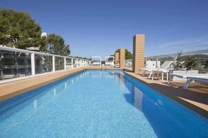Una piscina con muelle de madera, sillas de descanso y baranda de cristal.