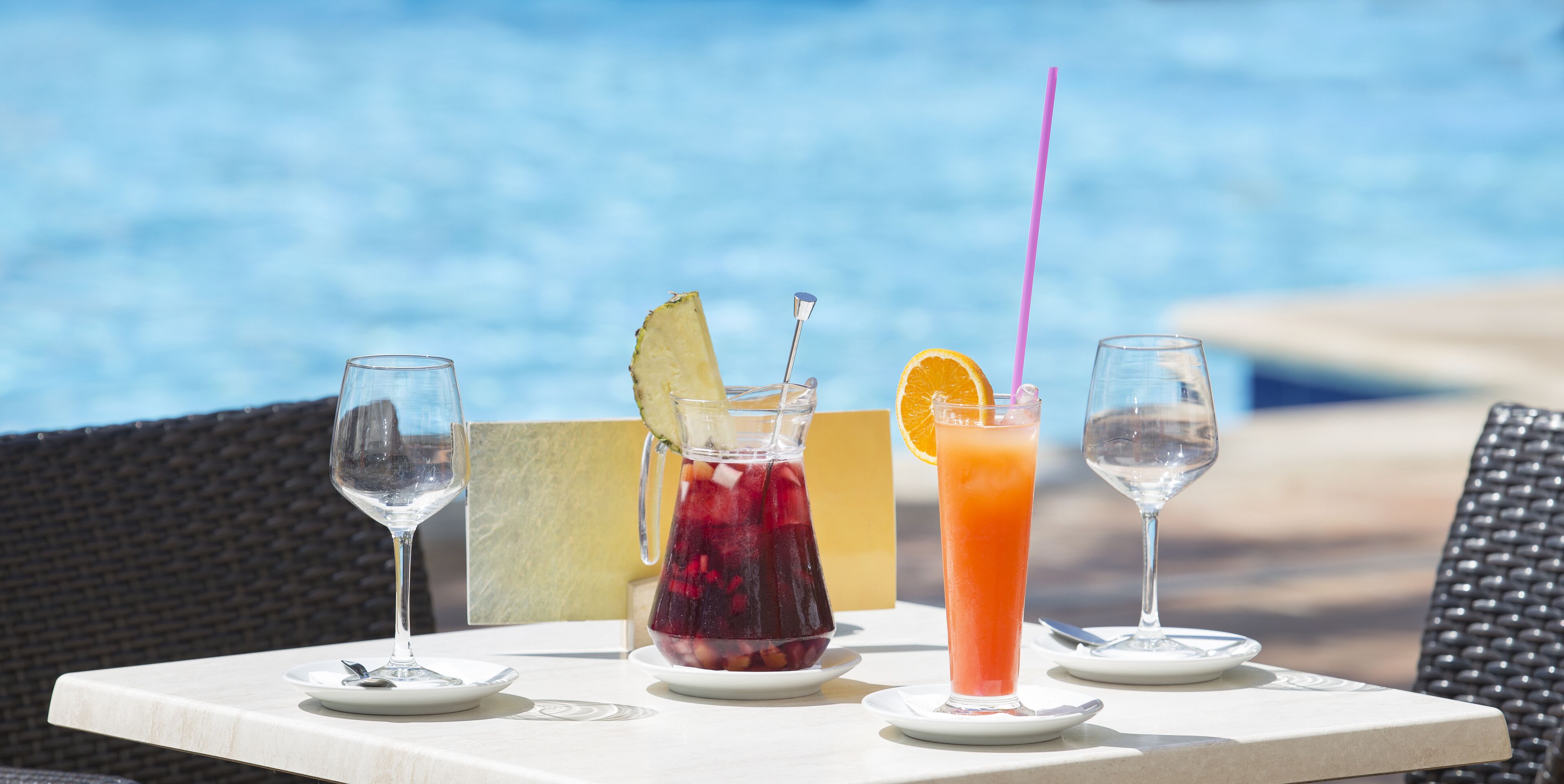 A table with four glasses of beverages, a pitcher, and a plate with a spoon, set against a poolside background.