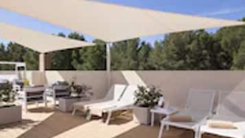 Un espacio de estar al aire libre cubierto con sillas reclinables blancas y una mesa, rodeado por una terraza de madera y un dosel grande.