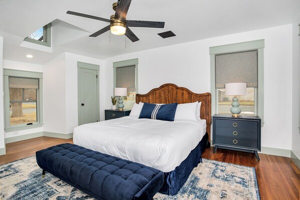 A bedroom with a large bed, a ceiling fan, two bedside tables with lamps, a bench, and a view of the outdoors through a window.