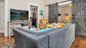 Uma sala de estar aconchegante com um sofá seccional cinza, uma televisão de tela plana e uma cozinha ao fundo.
