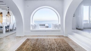 Un intérieur moderne avec une grande fenêtre en arc donnant sur la mer, un canapé blanc agrémenté de coussins et un tapis à motifs sur le sol.